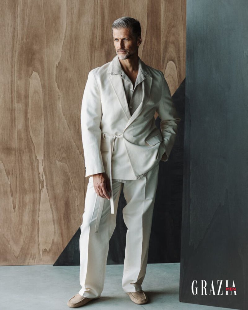 Mature man in an off-white suit poses against a geometric wooden backdrop, radiating confidence in a studio setting with Grazia logo.