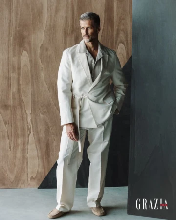 Mature man in an off-white suit poses against a geometric wooden backdrop, radiating confidence in a studio setting with Grazia logo.