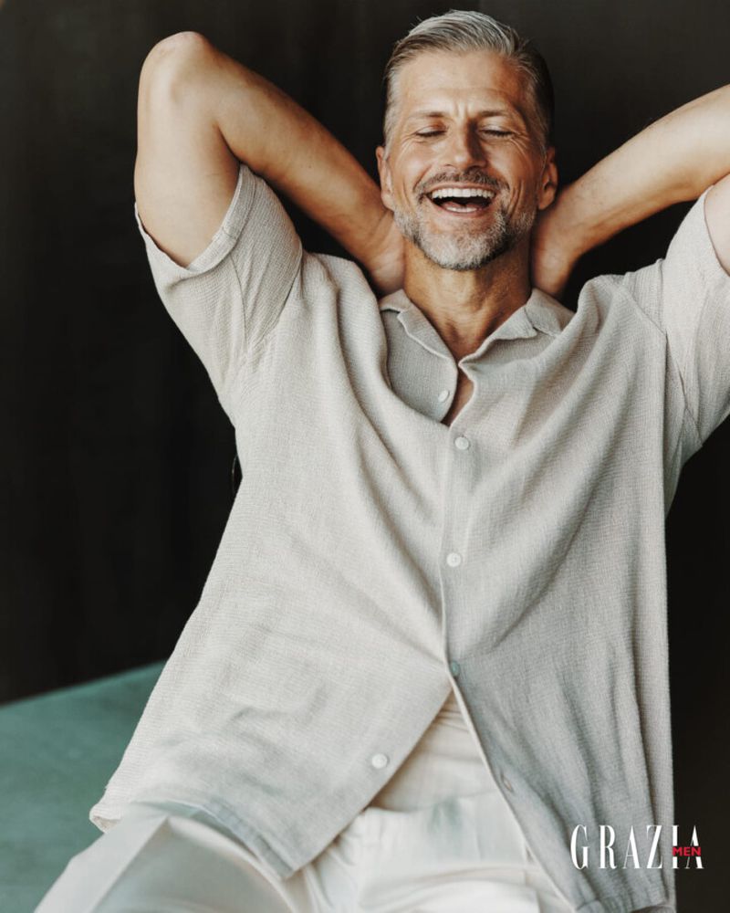 Smiling middle-aged man in a light-colored shirt poses with hands behind his head in a studio setting, fashion editorial style.