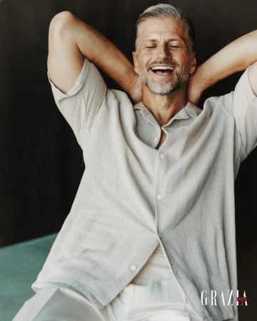 Smiling middle-aged man in a light-colored shirt poses with hands behind his head in a studio setting, fashion editorial style.