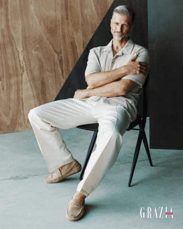 Model Frank Christians sits on a black chair wearing a beige camp shirt, white trousers, and tan suede loafers in a studio.