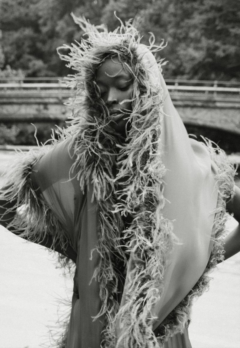 Black and white fashion portrait of a woman wearing a feathered shawl and veil in an outdoor location with a stone bridge in the background.