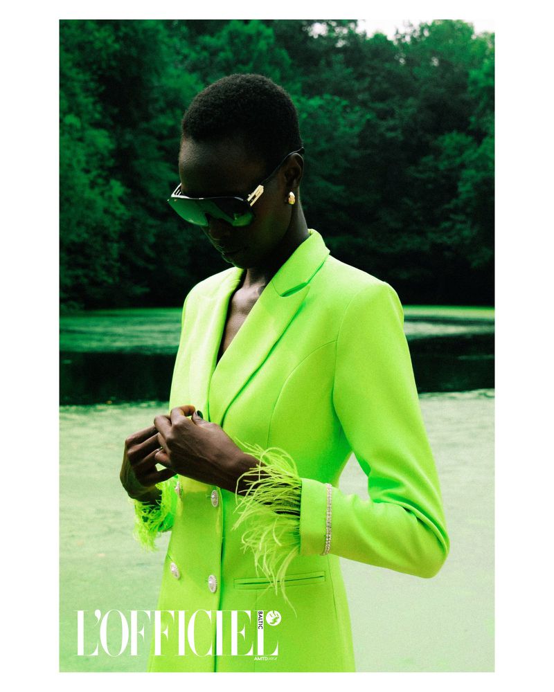 A slender Black woman with sunglasses poses in a neon green blazer with feathered sleeves outdoors against a blurred green forest background.