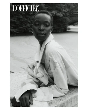 Black and white portrait of a Black woman with short hair leaning on a stone balustrade wearing a light trench coat outdoors.