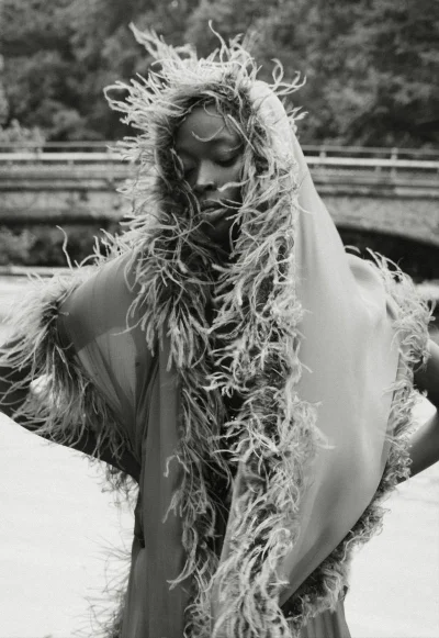 Black female model wearing an elaborate feathered hood and sheer shawl outdoors in a black and white portrait.