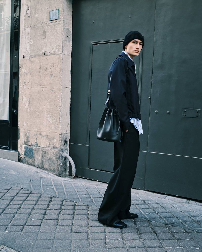 A male model in a dark jacket and wide-leg trousers poses against a gray door on a cobblestone street, wearing a black beanie and carrying a black bag.