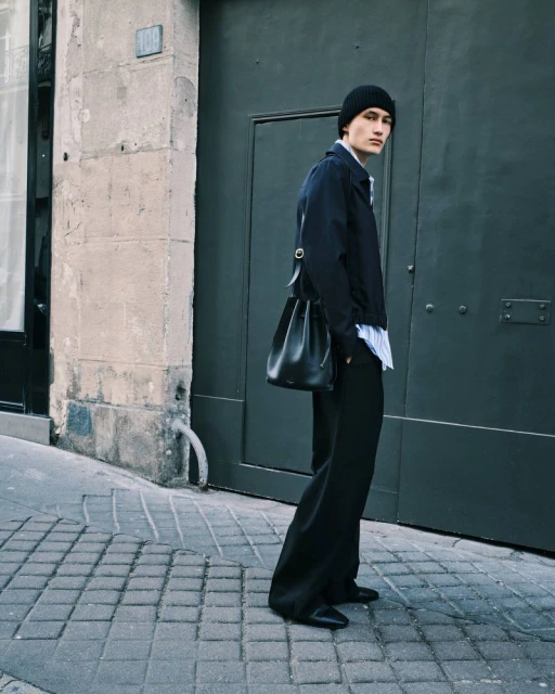 A male model in a dark jacket and wide-leg trousers poses against a gray door on a cobblestone street, wearing a black beanie and carrying a black bag.