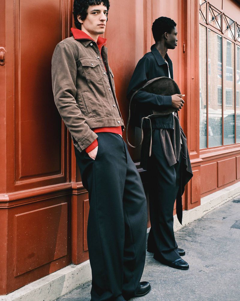 Two men pose in stylish outerwear on a city street, one in a brown corduroy jacket, the other in a dark jacket and black pants.