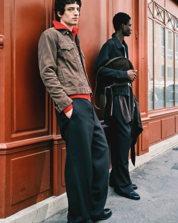 Two men pose in stylish outerwear on a city street, one in a brown corduroy jacket, the other in a dark jacket and black pants.