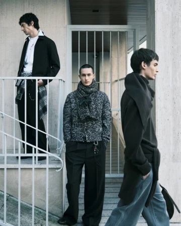 Three young adult men pose outdoors in fashionable dark-toned clothing with concrete and metal architectural elements in the background.