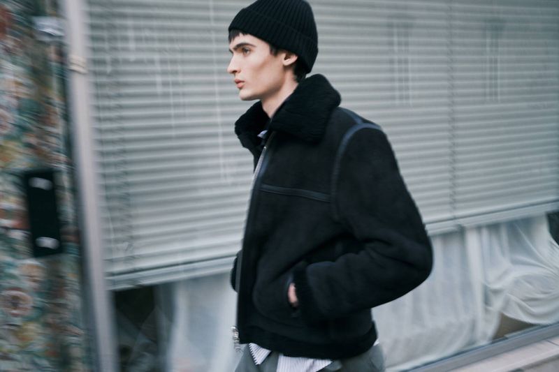 Man wearing a black jacket and beanie walking outdoors in front of a building with closed window shutters and a colorful wall.
