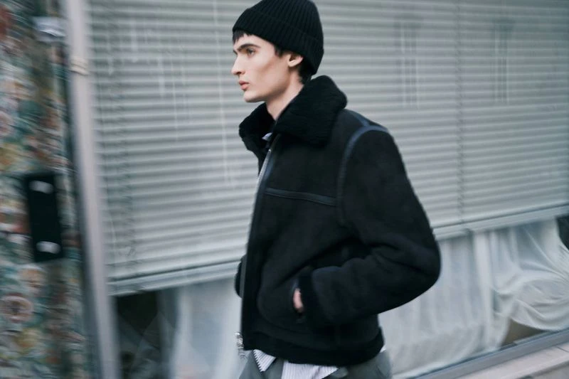 Man wearing a black jacket and beanie walking outdoors in front of a building with closed window shutters and a colorful wall.