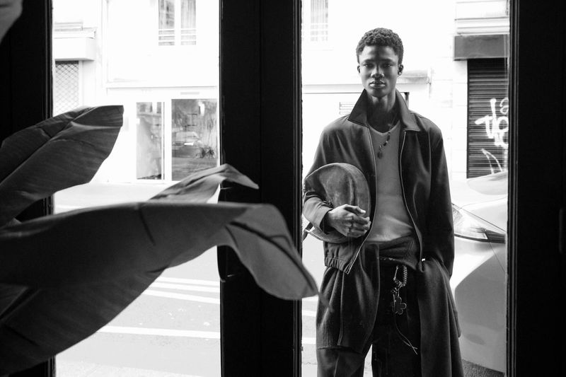 Black and white portrait of a Black male model in a jacket holding a curved bag, framed by a glass doorway.