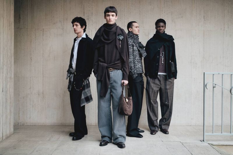 Four men stand in a line against a textured concrete wall. They are dressed in fashionable, casual clothing, accessorized with scarves and bags.