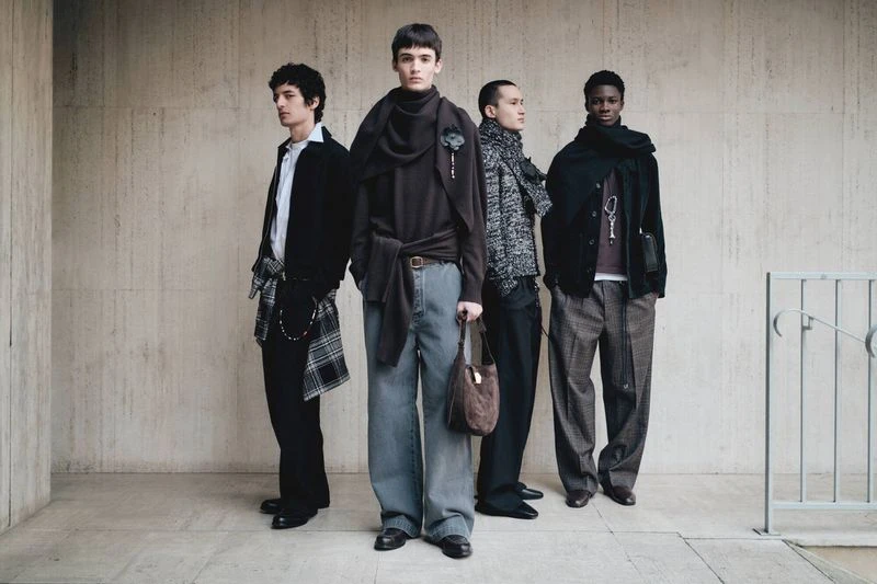 Four men stand in a line against a textured concrete wall. They are dressed in fashionable, casual clothing, accessorized with scarves and bags.