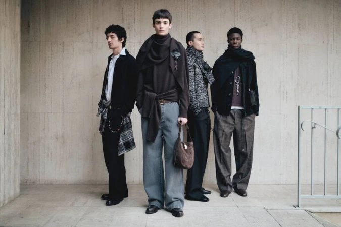 Four men stand in a line against a textured concrete wall. They are dressed in fashionable, casual clothing, accessorized with scarves and bags.