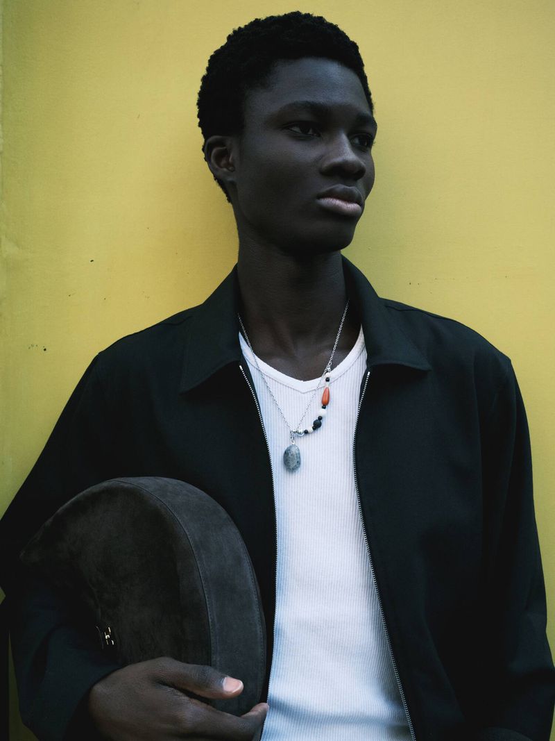 Dark-skinned man in casual jacket against yellow wall, holding drum. Silver necklace and white ribbed t-shirt visible in medium shot.
