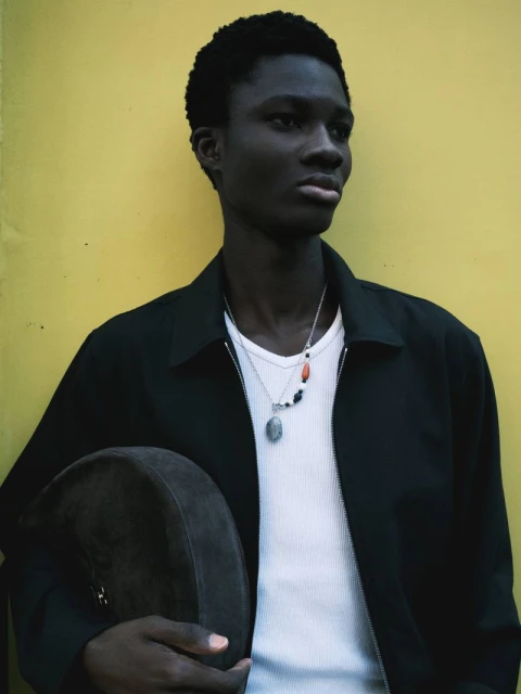 Dark-skinned man in casual jacket against yellow wall, holding drum. Silver necklace and white ribbed t-shirt visible in medium shot.