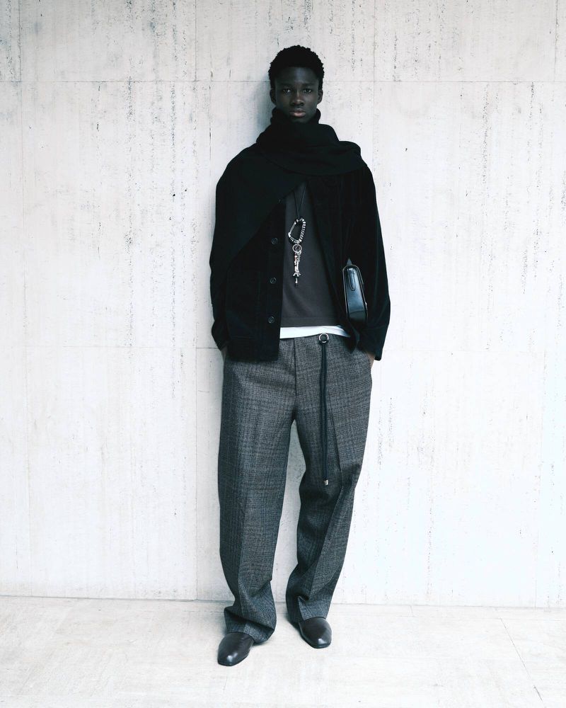 Dark-skinned adult male wearing a black jacket and grey patterned pants posing against a concrete wall in a full-length shot.
