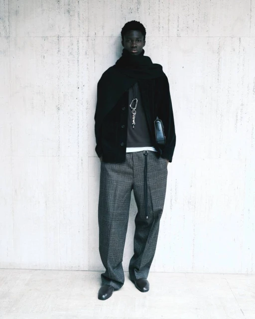 Dark-skinned adult male wearing a black jacket and grey patterned pants posing against a concrete wall in a full-length shot.