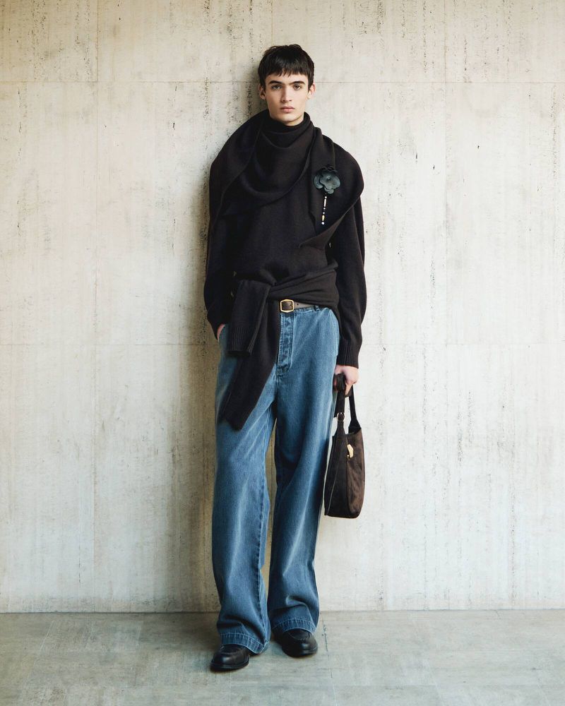 Full-length portrait of a male model wearing a black draped sweater and blue jeans, holding a small dark handbag against a textured concrete wall.