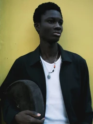 A Black male model wearing an open black jacket, white ribbed tank top, and layered necklaces against a solid yellow backdrop.