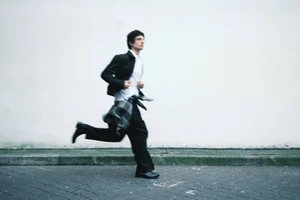 A young man running along a paved sidewalk past a white wall, wearing a black blazer, white shirt, and black trousers.
