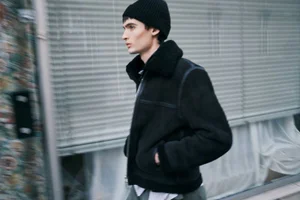 Young man in a black jacket and beanie walking past slatted blinds in an editorial portrait photograph.