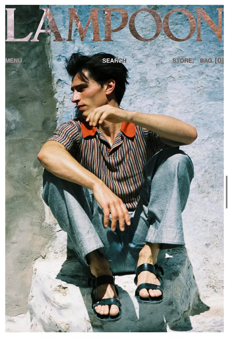 A young adult man sits on a stone structure wearing an orange and brown striped collared shirt, grey pants, and black sandals in daylight.