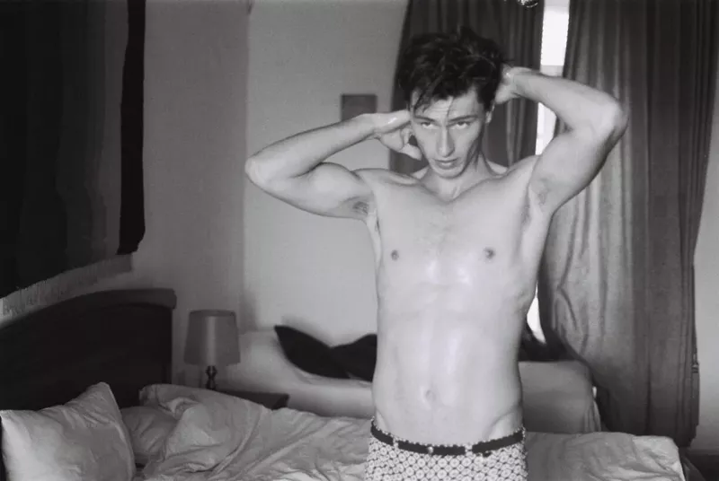 Black and white portrait of a shirtless adult male with hands behind his head, standing in a bedroom with a bed and window.