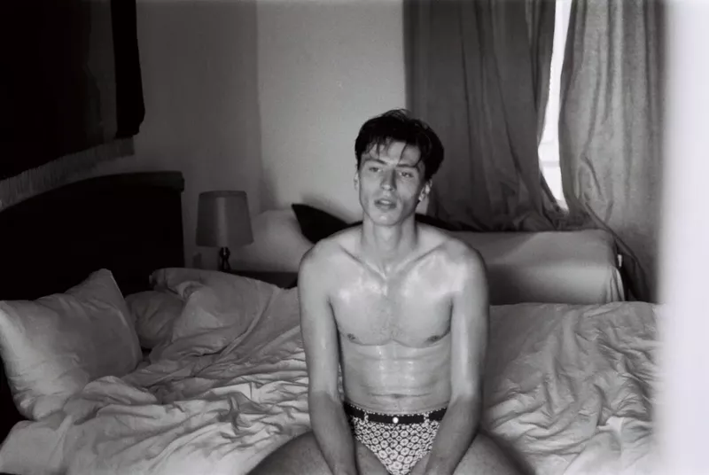 Black and white portrait of a shirtless adult male sitting on a bed with white sheets in an indoor bedroom setting.