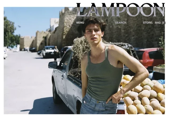 Adult male poses near a truck filled with fruit. The background features an old stone structure and other vehicles on the street.