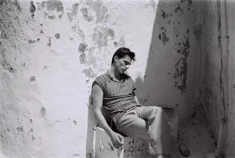 Monochrome portrait of a slender adult male with dark hair sitting in a room against a textured wall, lost in thought.