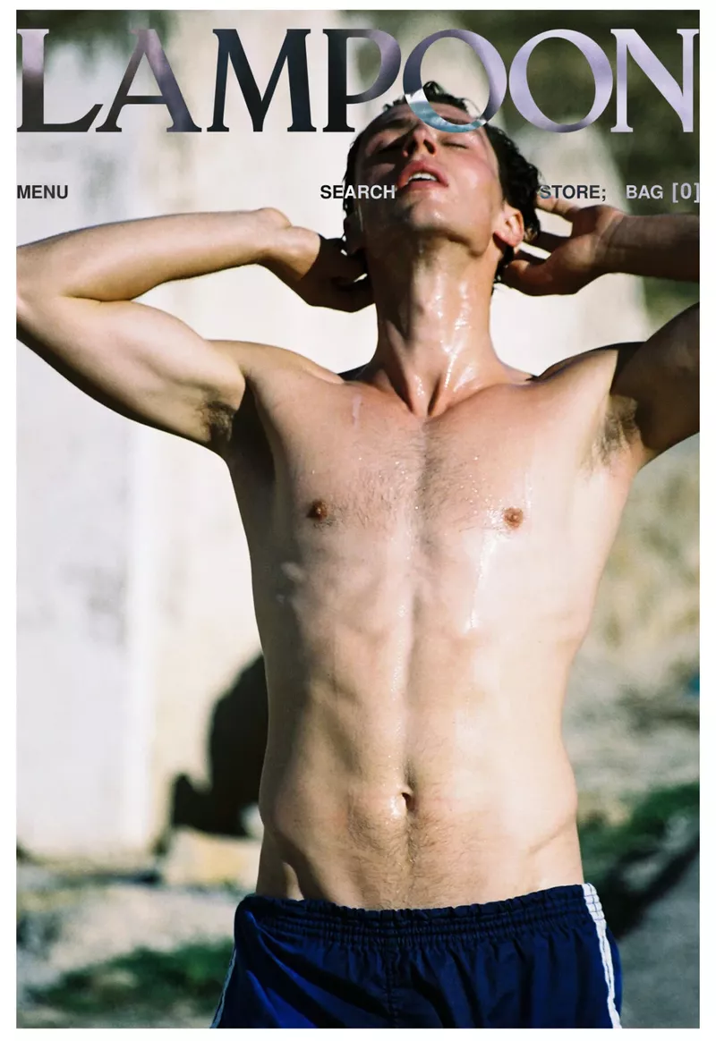 Shirtless man with blue athletic shorts poses with hands behind his head in front of a wall, in a fashion editorial shot.
