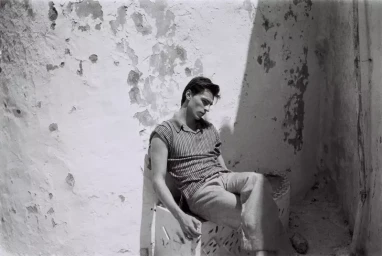 Male model seated against a weathered plaster wall wearing a striped polo and light trousers for Lampoon Magazine editorial