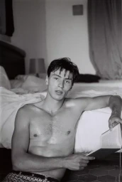 Monochrome portrait of a shirtless male model holding a book while seated on the floor in front of a bed
