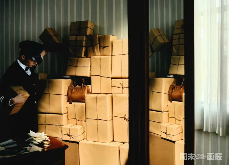 Man in uniform stacks wrapped parcels, a handbag placed among them, against a striped wallpaper backdrop in a dimly lit indoor setting.