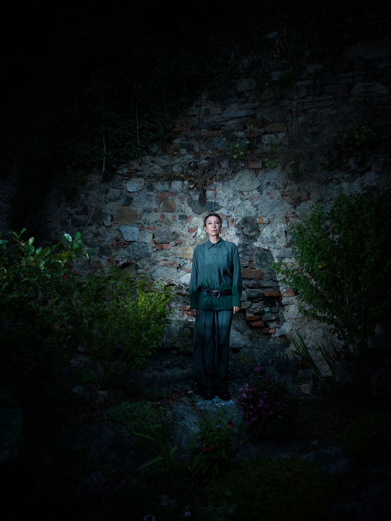 An adult female stands against a stone wall amidst greenery, bathed in dim, focused light creating a dramatic and moody outdoor portrait.