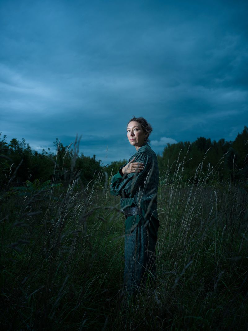 Woman stands in a field of tall grass beneath a cloudy sky. She is dressed in green clothing and has her arms crossed. Low-key lighting.