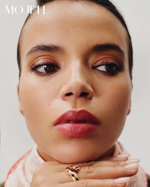 Close-up studio portrait of an adult female with hand under chin, wearing a gold ring, glossy red lipstick, and natural makeup.