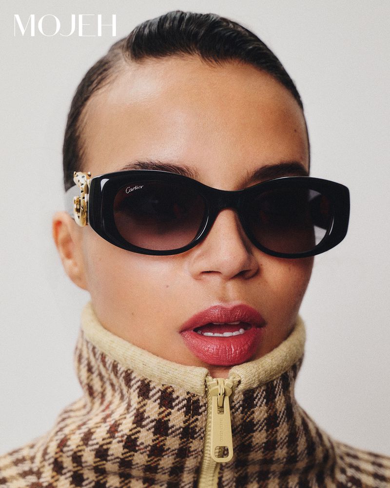 Glamorous woman in a houndstooth jacket and designer sunglasses poses in a studio, showcasing high-end fashion and beauty.
