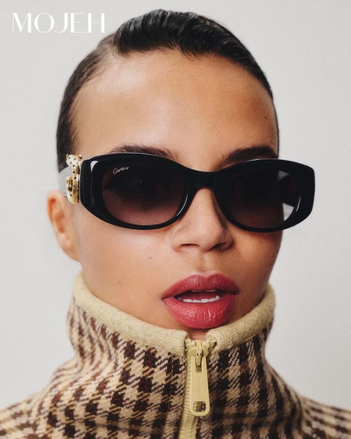Glamorous woman in a houndstooth jacket and designer sunglasses poses in a studio, showcasing high-end fashion and beauty.