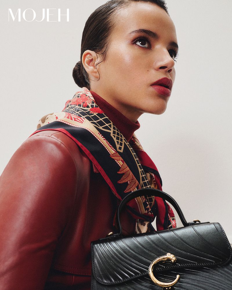 Woman wearing a red jacket and patterned headscarf holding a black handbag. Close up, looking up, with a white background.