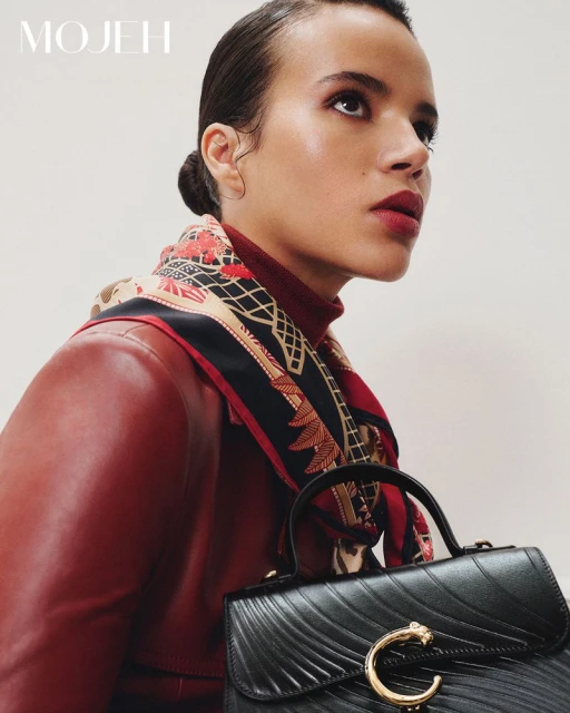 Woman wearing a red jacket and patterned headscarf holding a black handbag. Close up, looking up, with a white background.