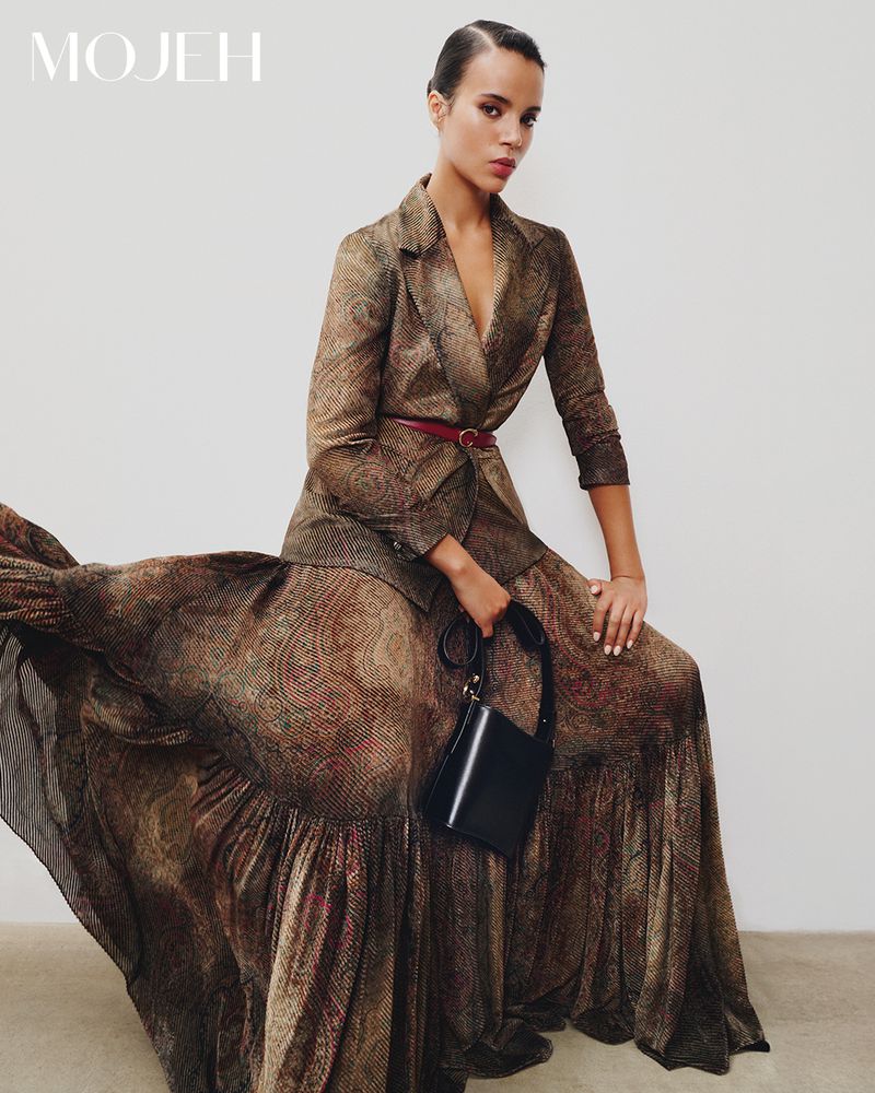 An adult female model wears a patterned brown blazer and matching flowing skirt, accessorized with a thin belt and black purse against a plain background.