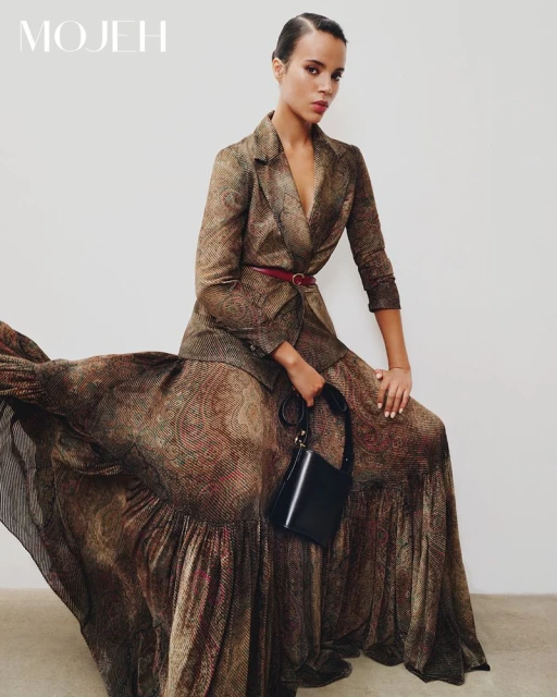 An adult female model wears a patterned brown blazer and matching flowing skirt, accessorized with a thin belt and black purse against a plain background.