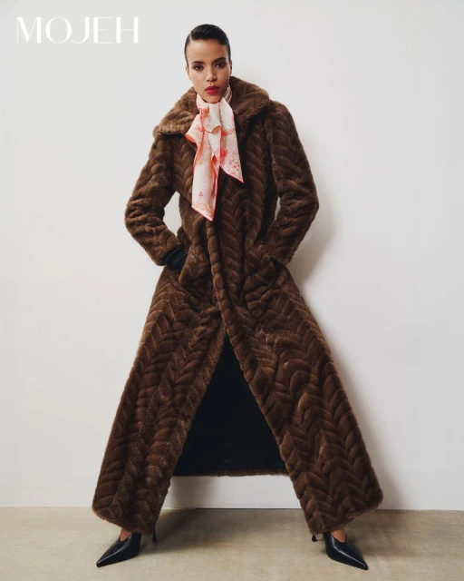 Full-length shot of an adult woman in a brown fur coat with a silk scarf, posing against a white wall in a fashion editorial.