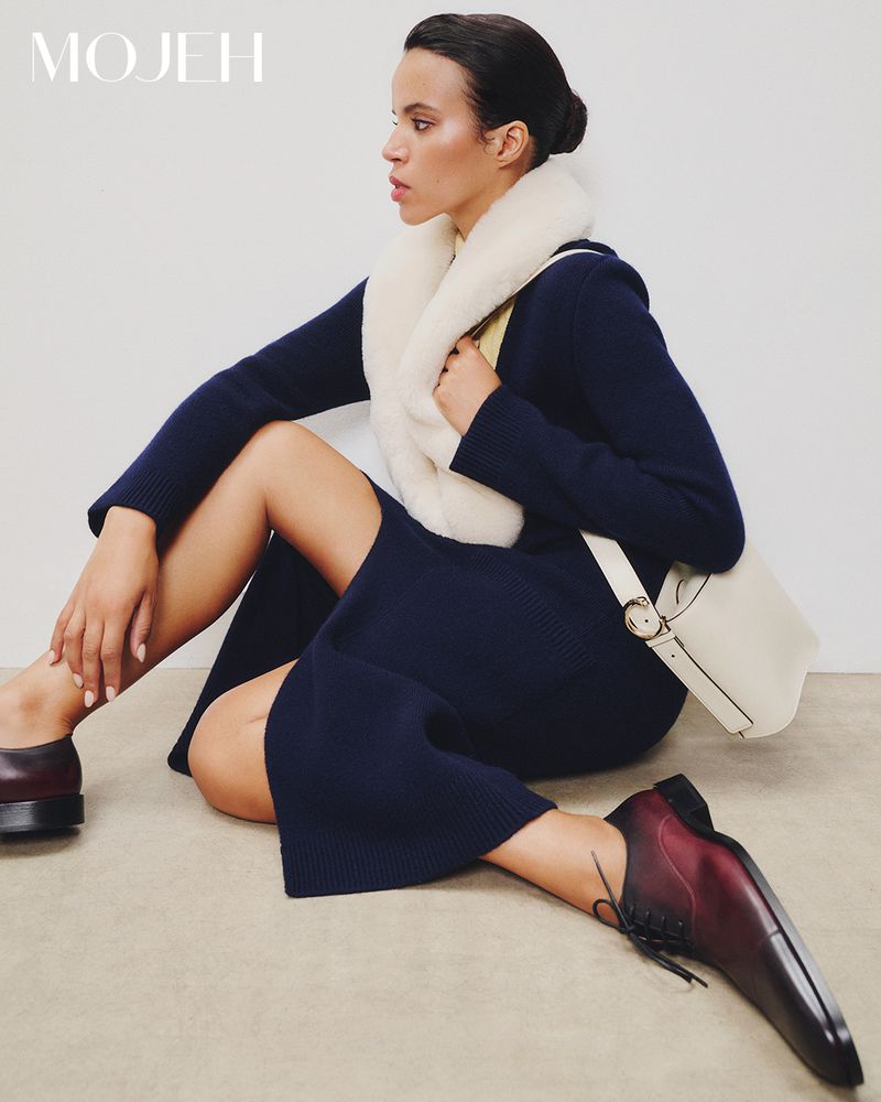 A woman in a dark dress and burgundy shoes poses in a studio, wearing a white fur collar and carrying a white purse.