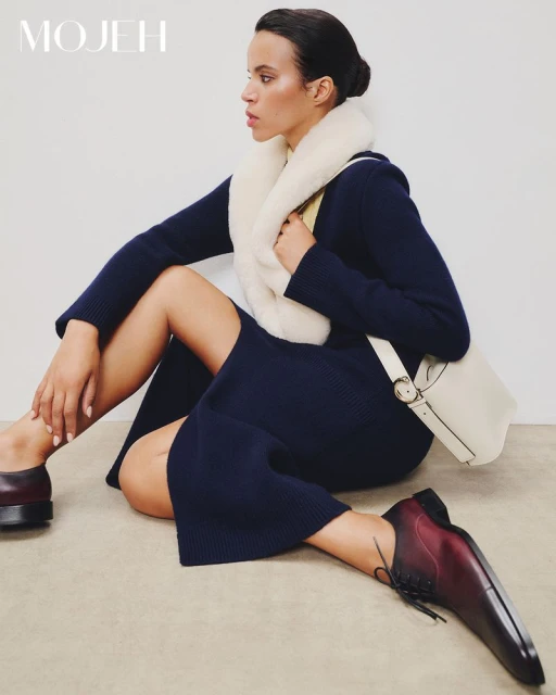 A woman in a dark dress and burgundy shoes poses in a studio, wearing a white fur collar and carrying a white purse.