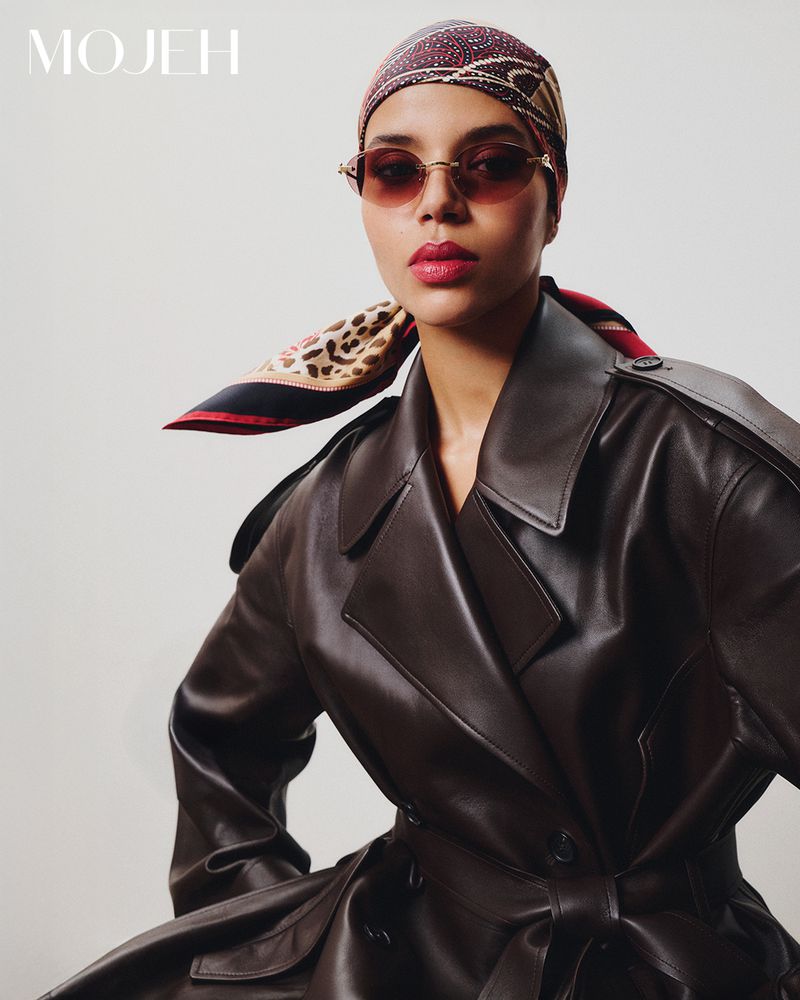A woman in a brown leather trench coat, patterned headscarf, and oval sunglasses poses against a pale backdrop in an indoor fashion shot.